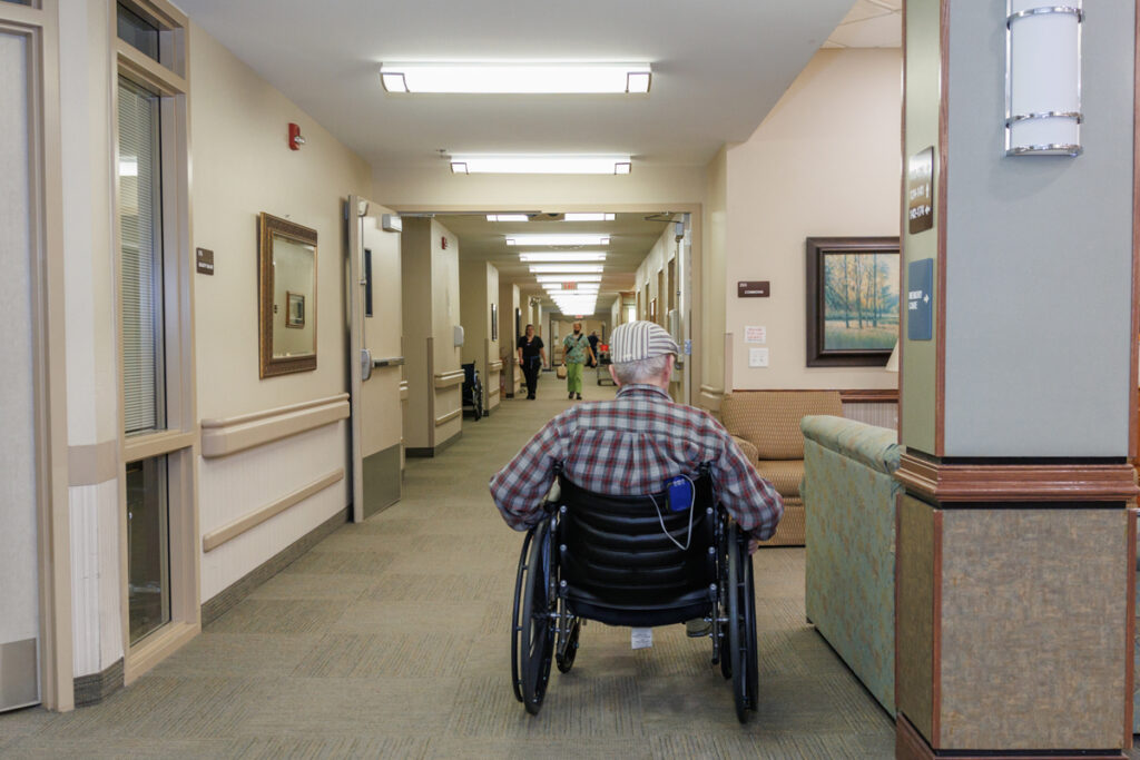 Resident using a wheelchair in a hallway at Westhaven Community in Boone, Iowa with handrails, seating area, and staff in the background.