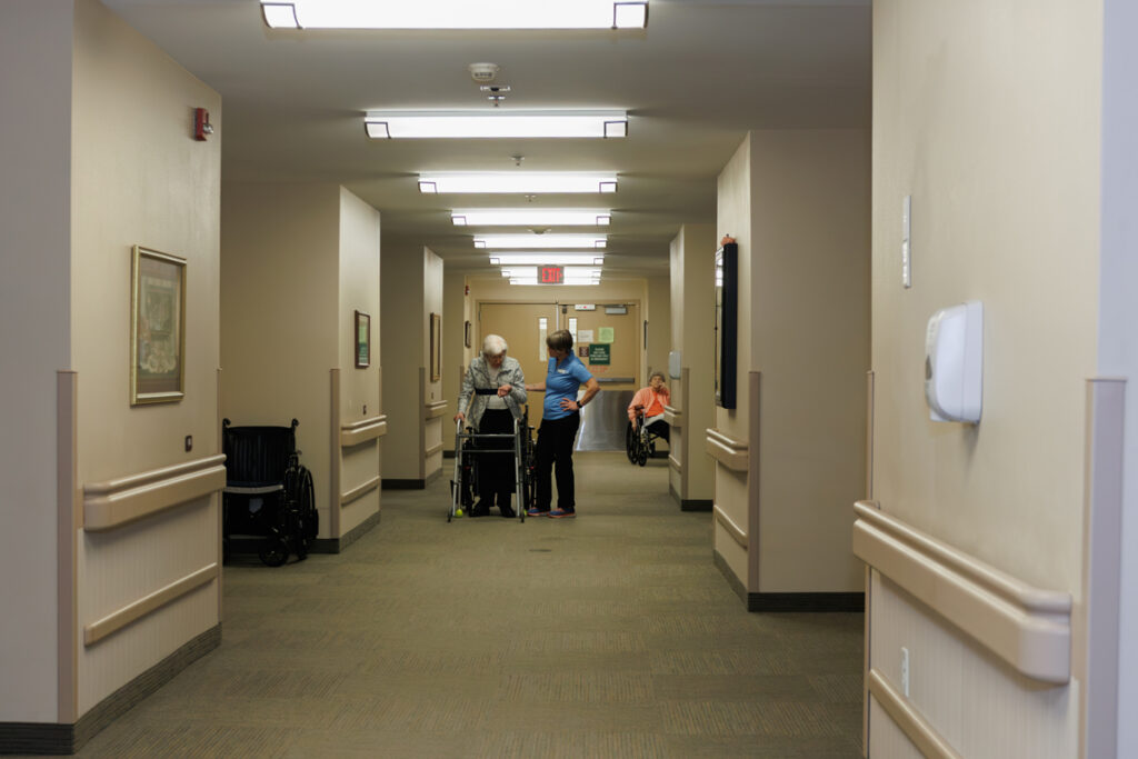 Hallway at Westhaven Community in Boone, Iowa with handrails, overhead lighting, and staff assisting a resident using a walker.